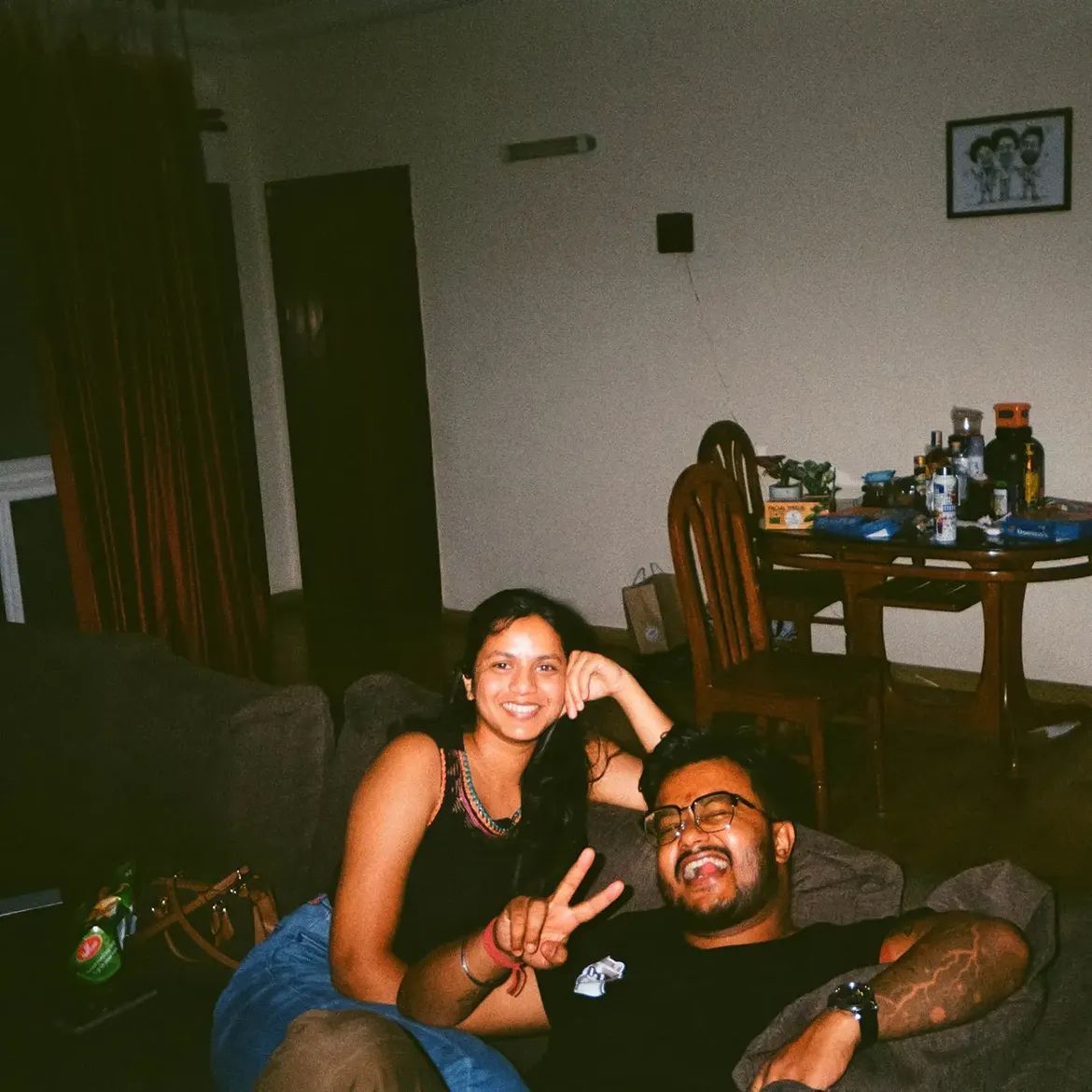 Two people on a sofa in a dim room, one resting an elbow on the other, both grinning at the camera.