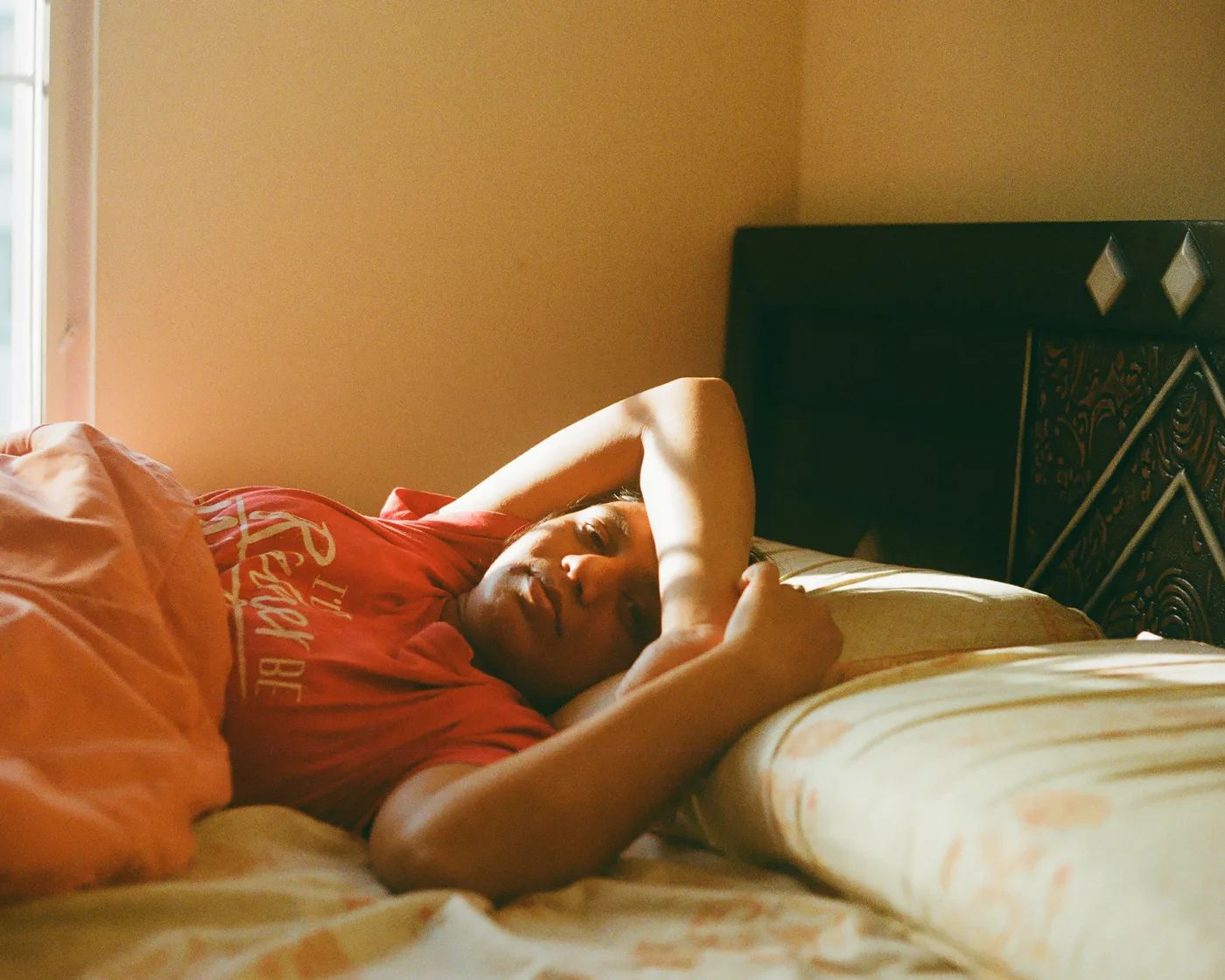 A person resting on a bed in a red t-shirt, late afternoon light coming in from a window beside them.