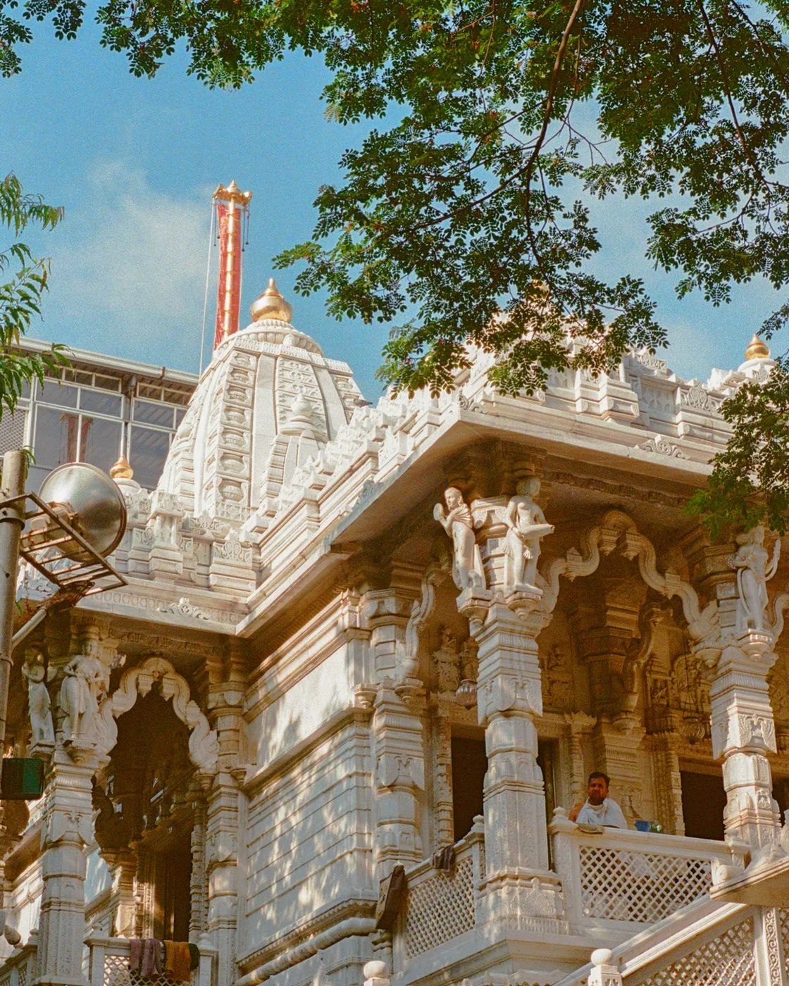 A carved white marble temple under a blue sky, seen through the branches of a tree, with a man standing on the balcony.