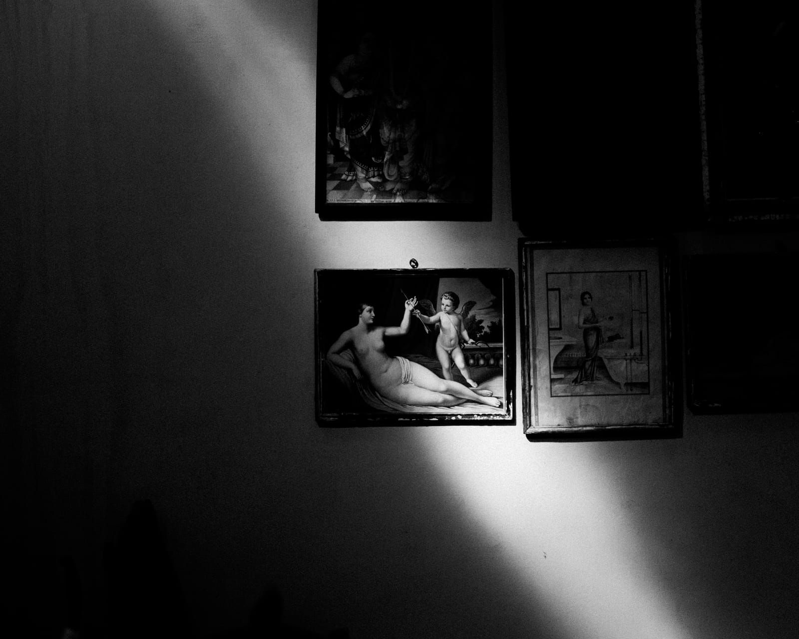 A shaft of daylight crossing a wall of old framed pictures in a dark room, in black and white.