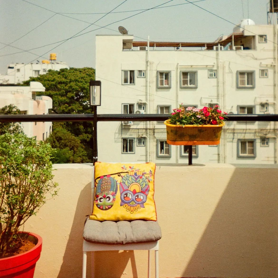 A cushion embroidered with two owls, on a chair on a balcony, flowers and apartment blocks behind.