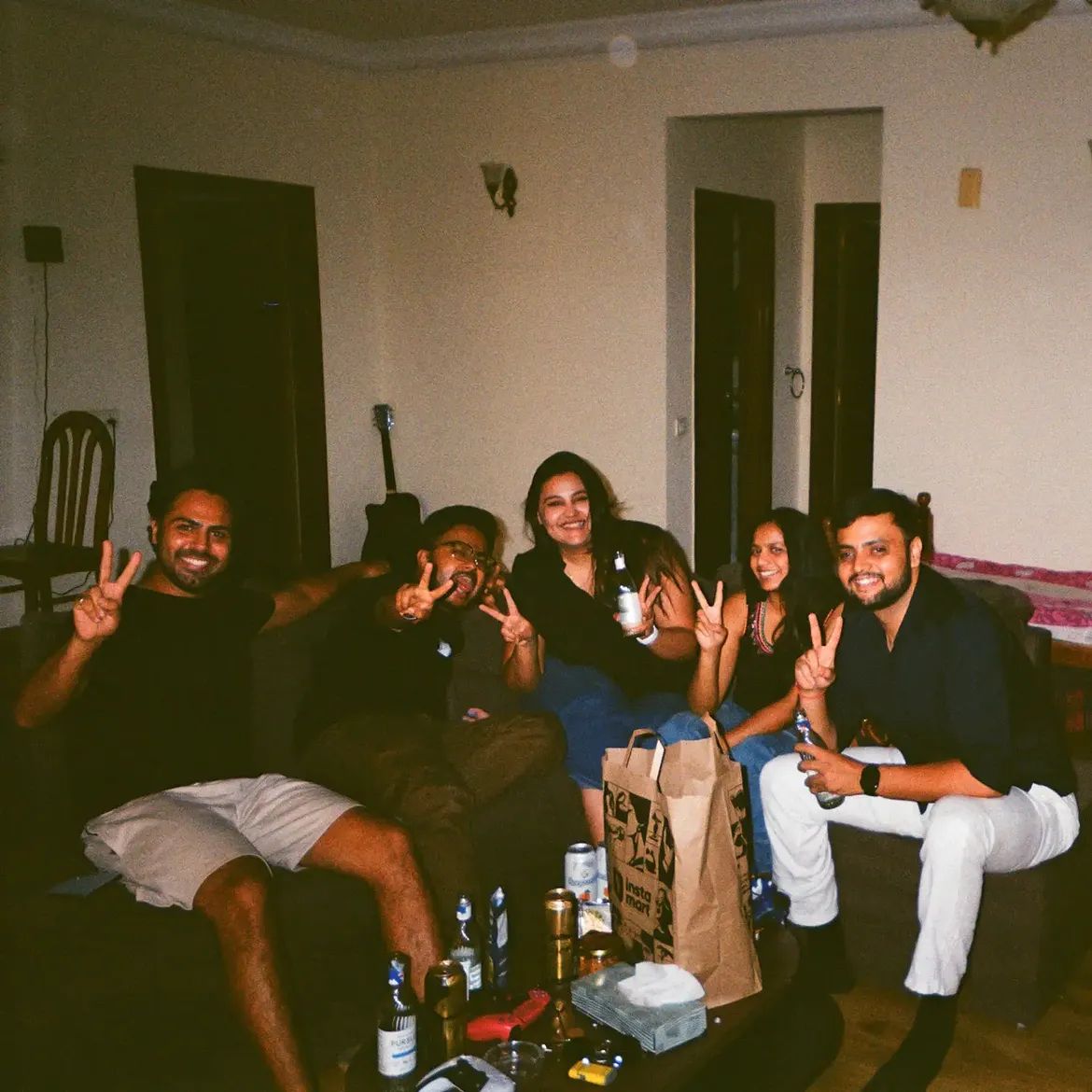 Five people crowded onto a sofa holding up peace signs, bottles and cans on the table in front.