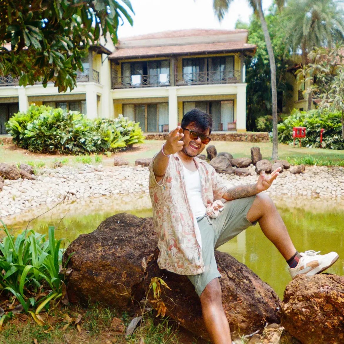 A person sitting on a boulder beside a green pond, arms out, a low tiled-roof building behind.