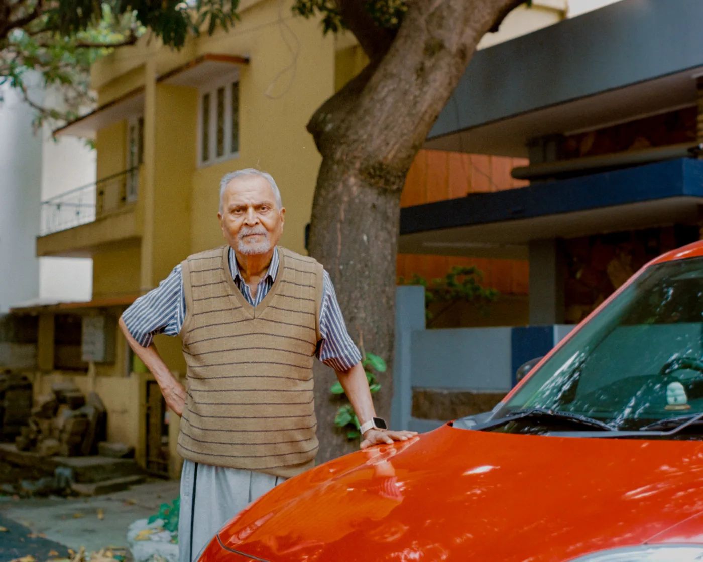 An older man standing with one hand resting on the bonnet of a red car, houses and a tree behind him.