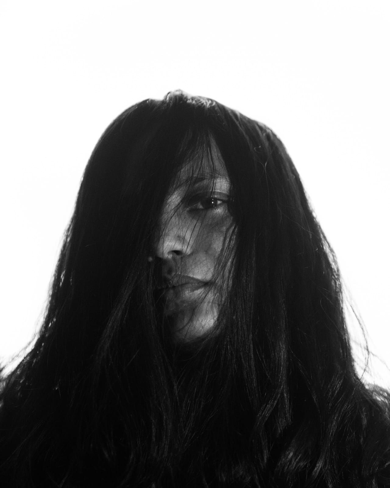 A black and white portrait against a white ground, long dark hair falling across most of the face.
