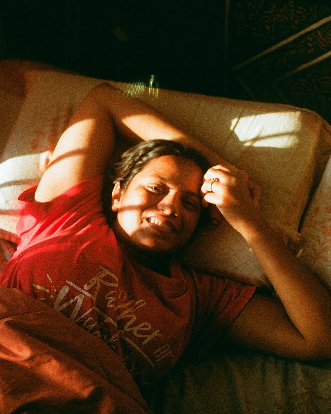 A person lying on a bed in a red t-shirt, grinning, with a band of sunlight falling across their face.