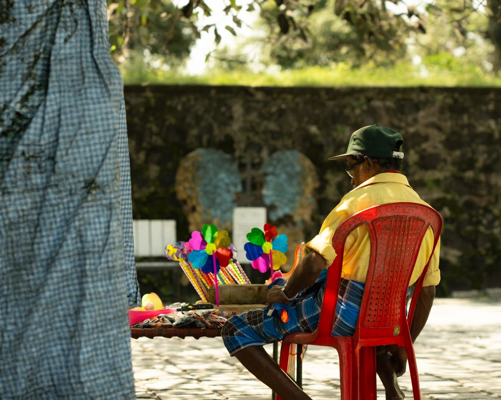 A street vendor sitting in a red plastic chair beside a stall of coloured paper pinwheels.