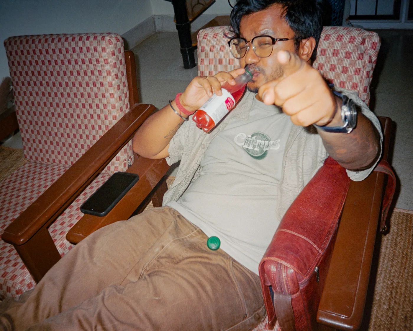 A man sprawled in a checked armchair drinking from a bottle, pointing at the camera.