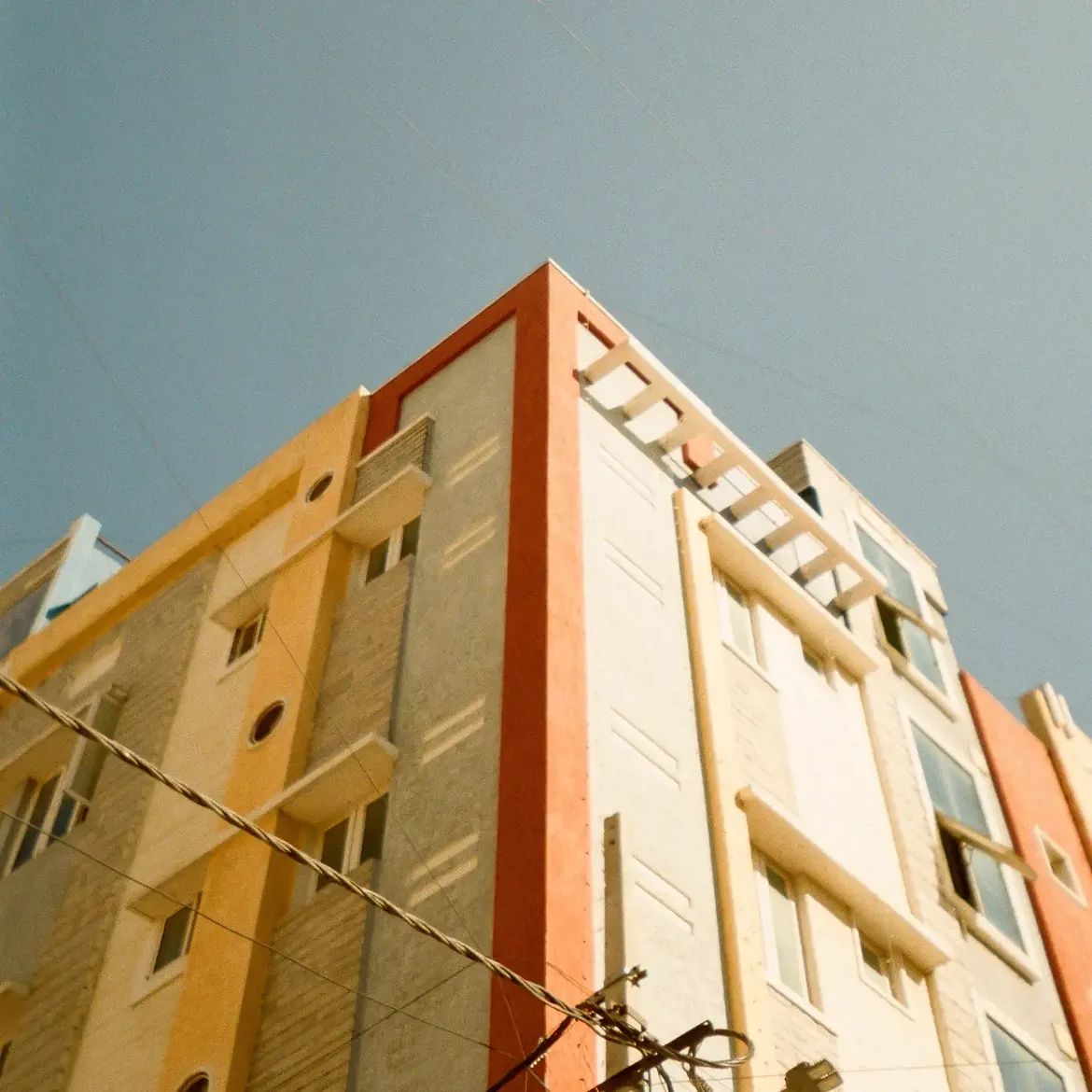 The corner of a painted apartment block shot from below against a clear sky, power lines crossing the frame.
