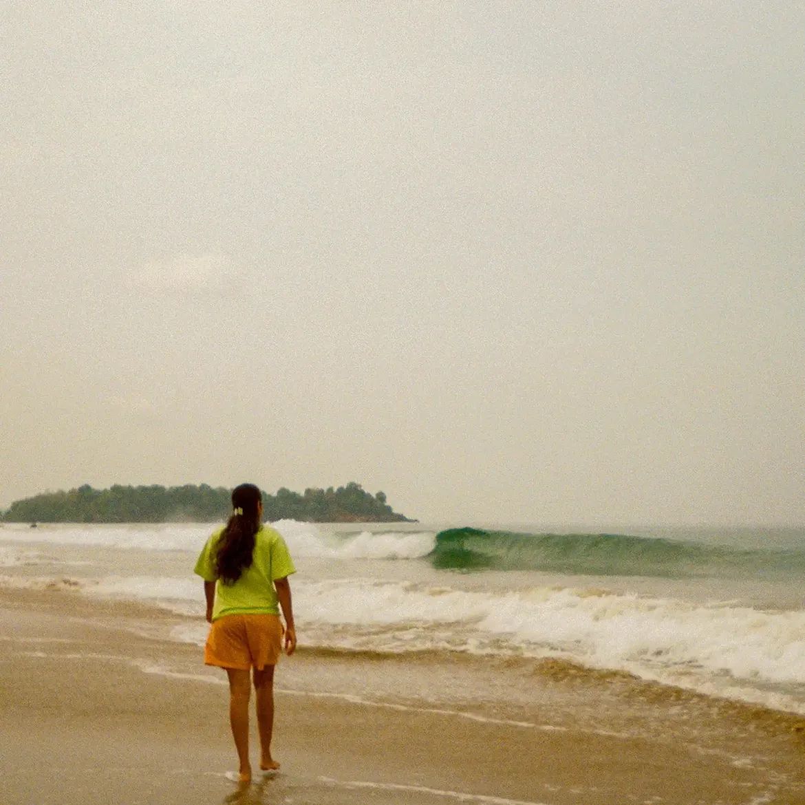 A person in a yellow shirt walking towards a breaking wave, a wooded headland in the haze behind.