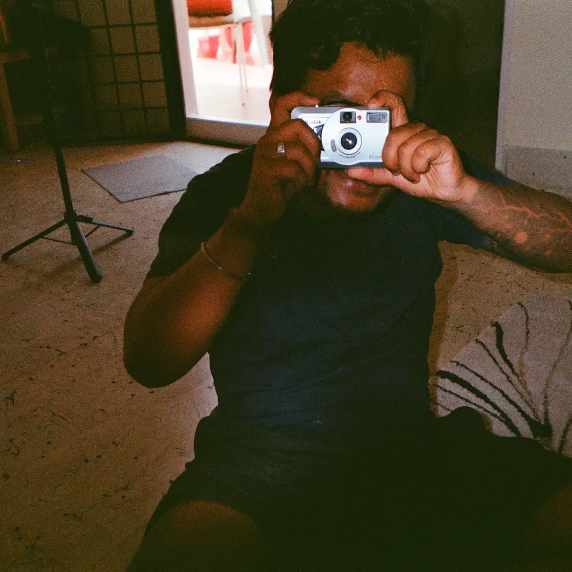 A person on the floor holding a silver Kodak KB10 up to their eye, pointing it back at the camera.
