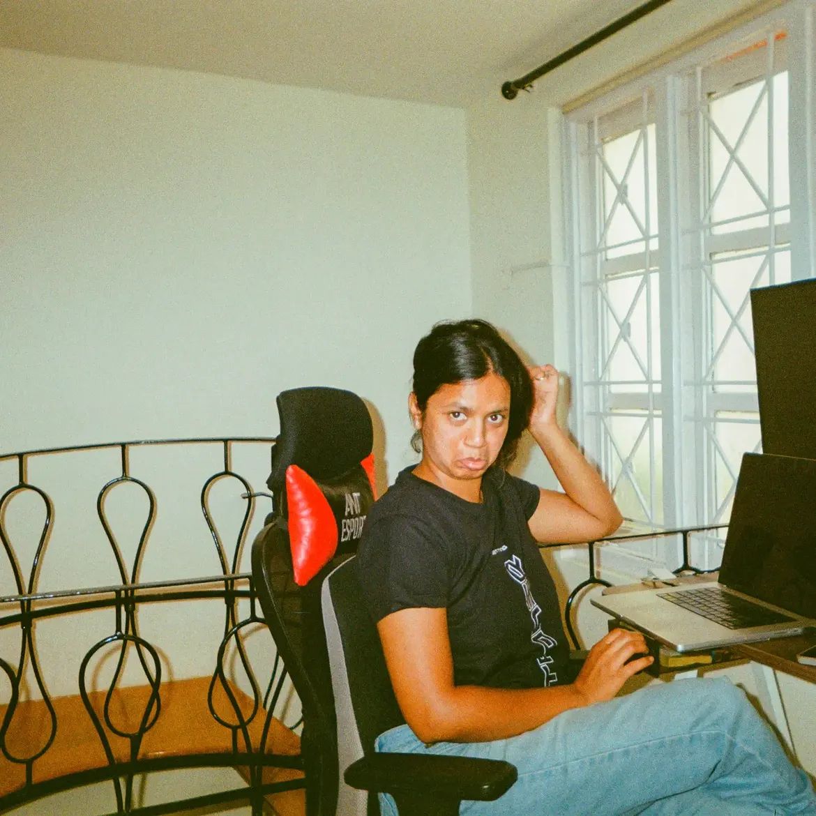 A person at a desk beside a tall window, turning away from the laptop to look at the camera.