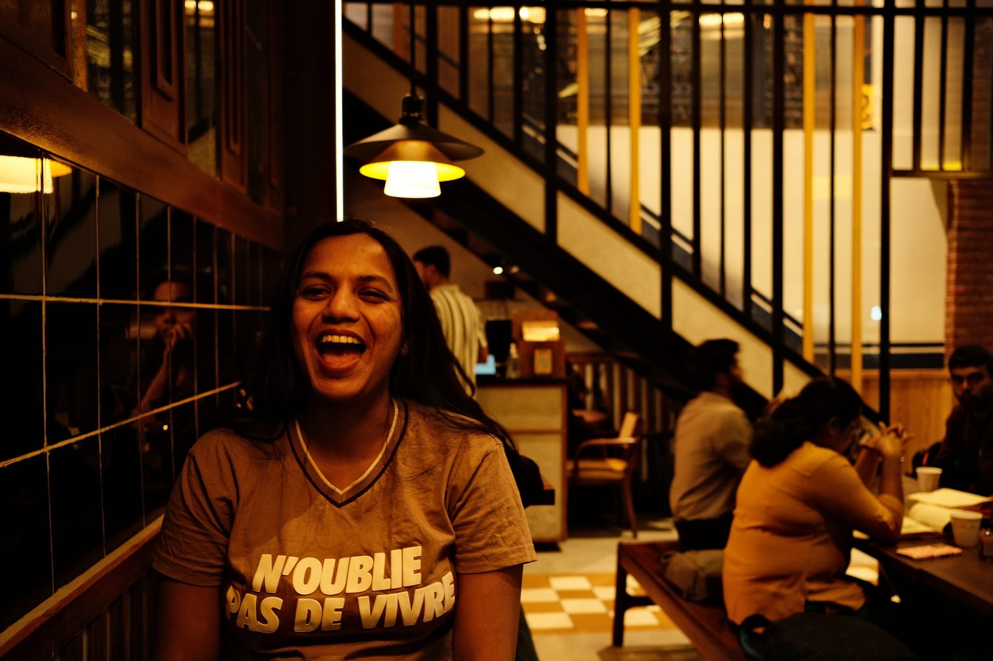 A woman laughing at the camera in a warmly lit café, in a t-shirt that reads N’oublie pas de vivre, with a staircase and other tables behind her.